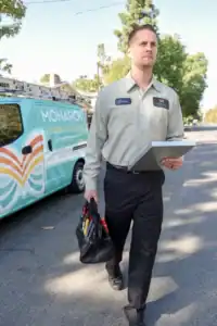 A technician in a uniform carries a tool bag and clipboard while walking on a street near a Monarch Home Services van, showcasing the rewarding careers and jobs available for those interested in joining the team.