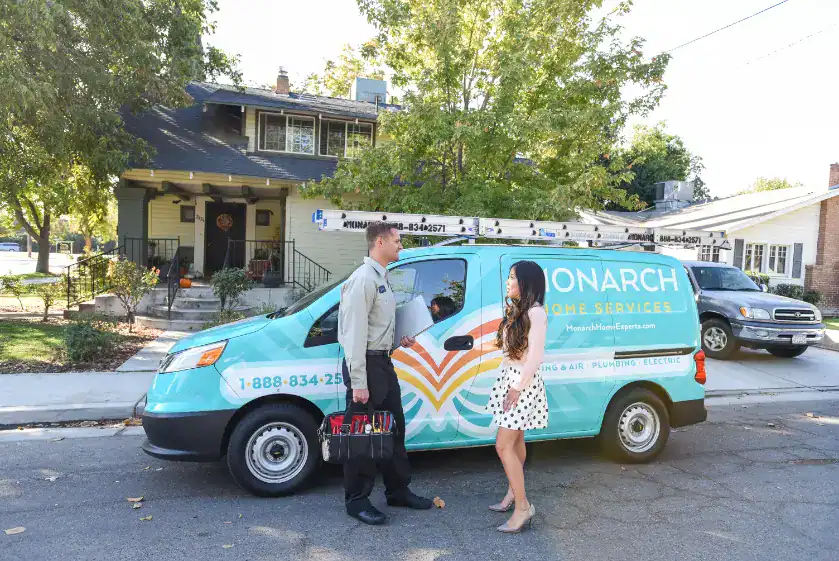 A technician holding a tool bag talks with a woman in front of a Monarch Home Services van parked on a residential street, demonstrating exceptional customer service.