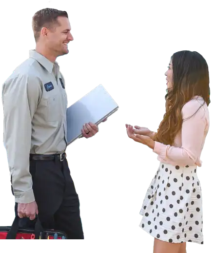 A man in a uniform holding a clipboard and tool bag talks with a woman in a pink blouse and polka dot skirt.