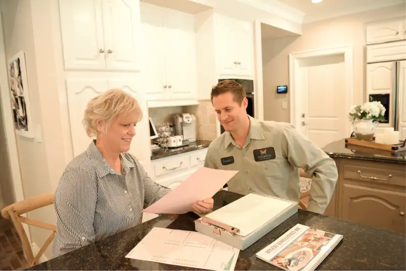 An older woman and a man in uniform review paperwork together at a kitchen counter lined with folders and a calendar.