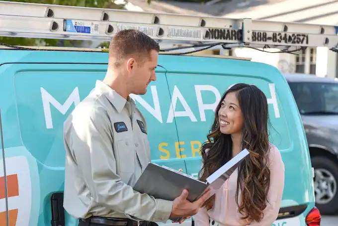 A technician holding a folder speaks with a woman in front of a van featuring “Monarch” branding, a mounted ladder, and clear contact information displayed on the side.